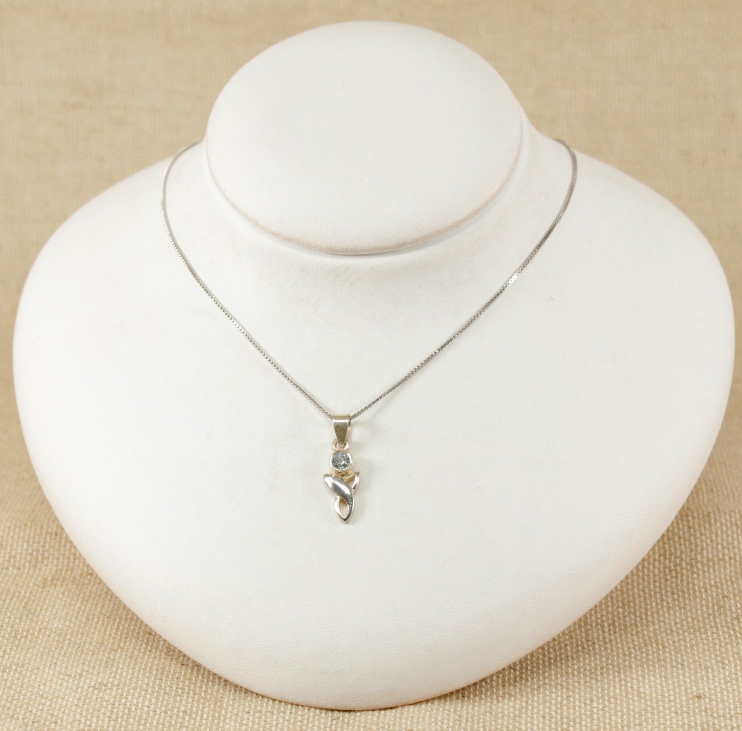 A silver pendant necklace with a round blue topaz set in a Celtic knot design, displayed on a white mannequin.