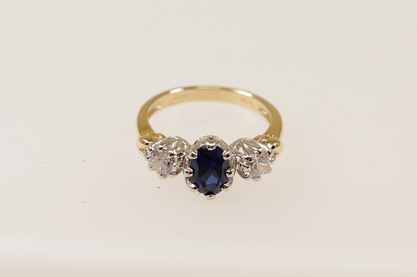 A vintage 9ct gold trilogy ring with a central blue oval-shaped sapphire gemstone and two smaller round cubic zirconia gemstones on either side, set against a white background.