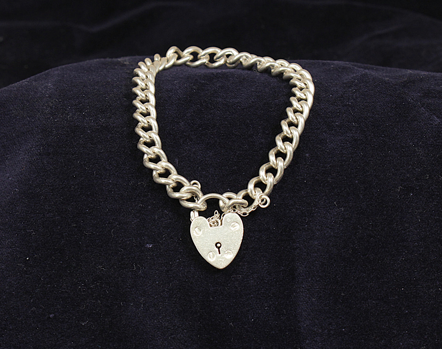 A silver curb link bracelet with a padlock fastening displayed on a dark fabric surface.