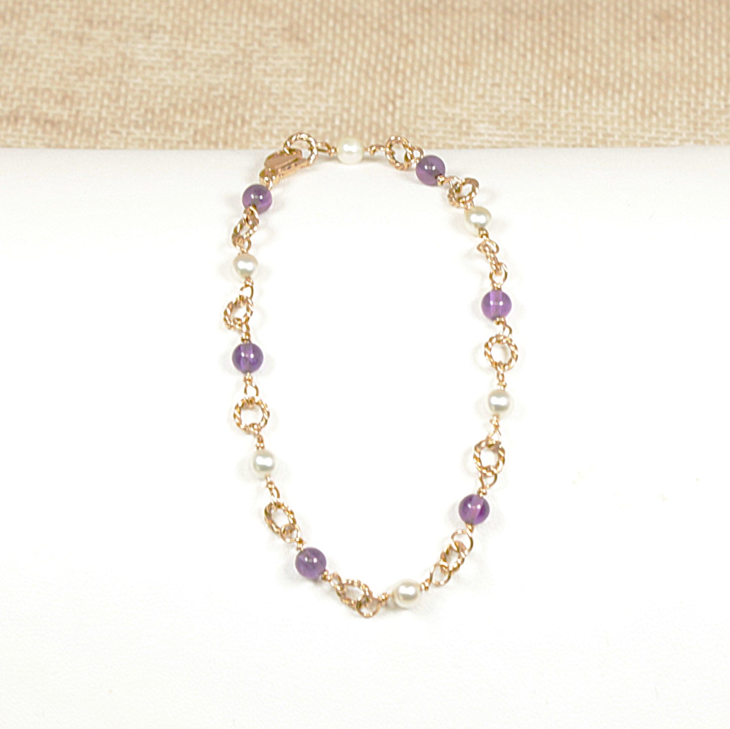 A 9ct gold bracelet with round cabochon amethyst stones and matching cultured pearls, featuring a fine gold rope threading and a gold lobster clasp, displayed against a beige background.
