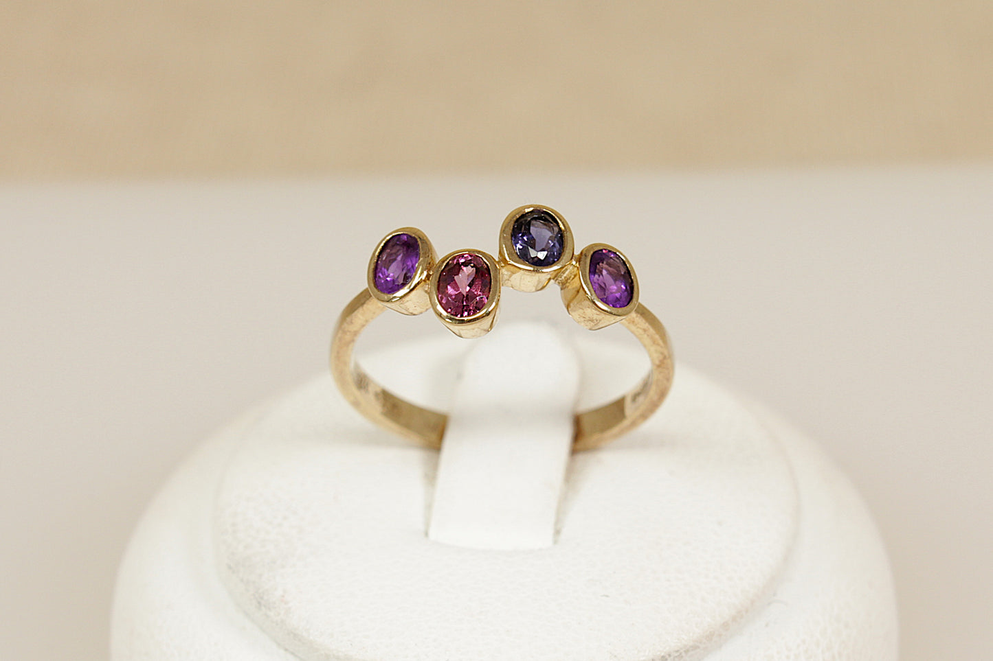 Gold multi gemstone ring with amethyst garnet and blue iolite gemstones on a white stand against a beige background