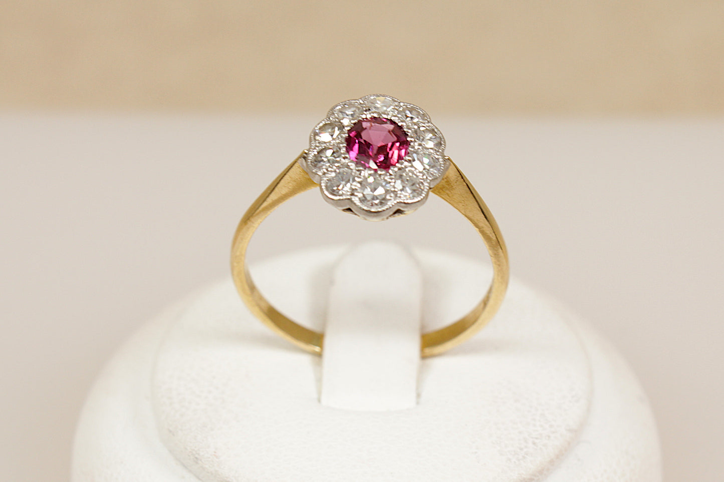 18ct ruby and diamond Gold ring with a halo of sparkling diamonds in a platinum mount on a white stand against a beige background