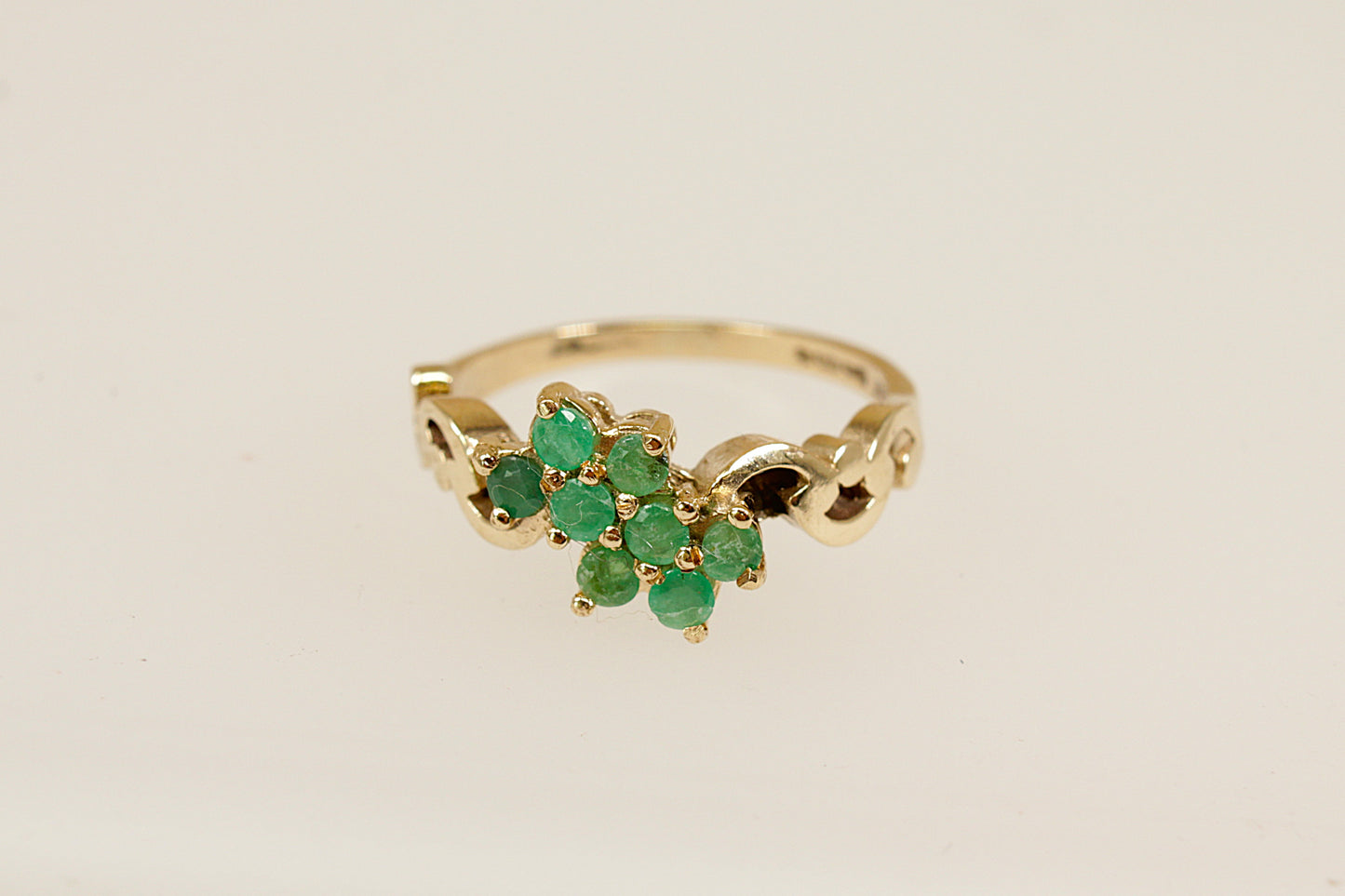 A charming vintage cluster ring crafted in warm 9ct yellow gold, featuring eight round-cut emeralds arranged in a delicate floral design. Each emerald is securely claw-set, creating a vibrant cluster that radiates natural green beauty. The ring's decorative shoulders are adorned with elegant scrolling goldwork, adding a touch of ornate detail and vintage character. Inside the band are full London hallmarks, with the date letter for 1979 and the maker’s mark ‘R & W’. UK finger size N.