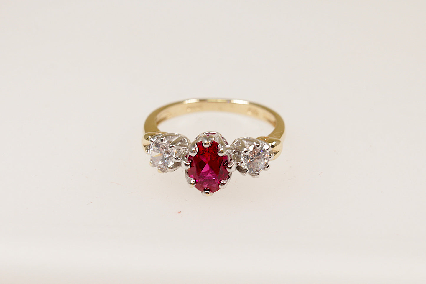 A 9ct gold trilogy ring with an oval cut ruby centerpiece and two round cut crystal gemstones on either side, set in a crown-inspired setting.