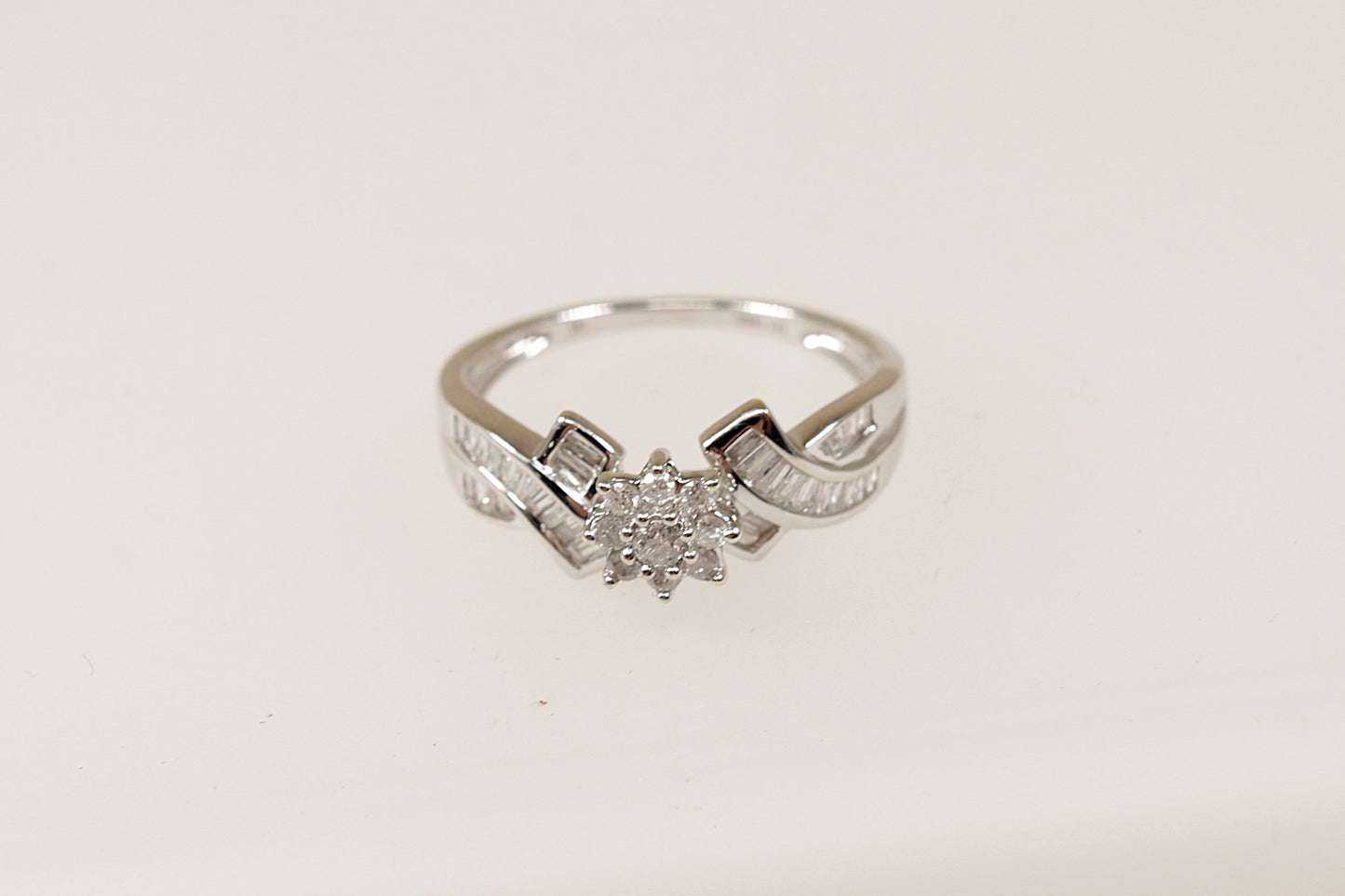 A white gold diamond cluster ring with a floral-inspired design, featuring a flower-shaped crown setting with nine petite diamonds, and baguette-cut diamonds on the shoulders.