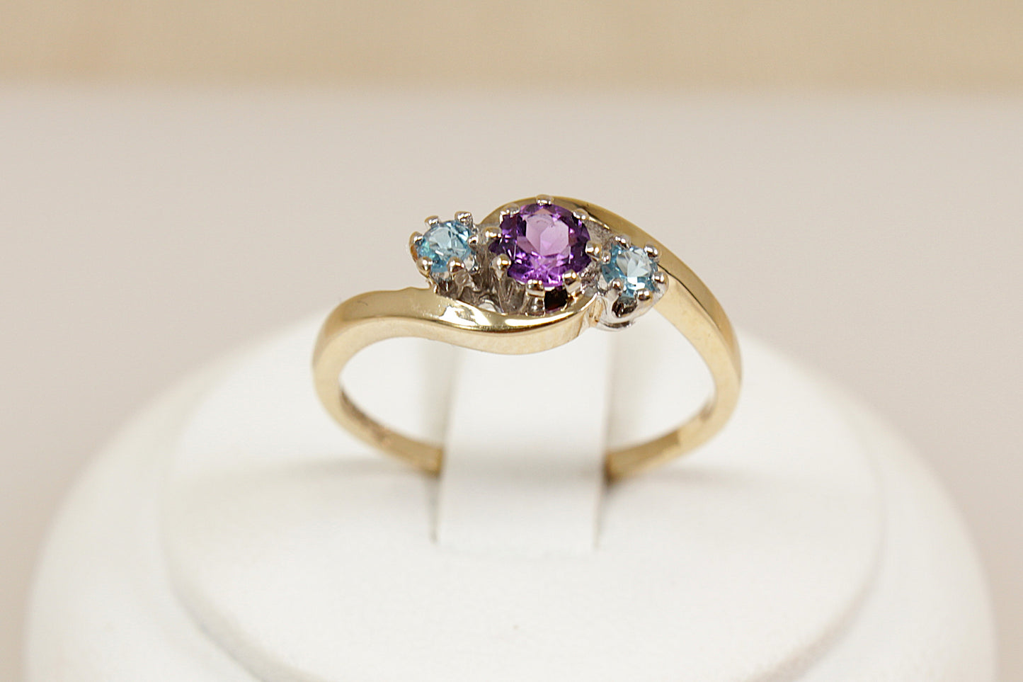A delightful and feminine 9ct gold trilogy ring, featuring a round claw-set Amethyst at the centre, flanked by two sparkling aquamarines on either side. The gemstones are elegantly arranged in a twist setting, giving the design a graceful flow and a touch of vintage romance, The ring is hallmarked in London for 1994, confirming its gold purity and vintage authenticity, UK Finger size: O.