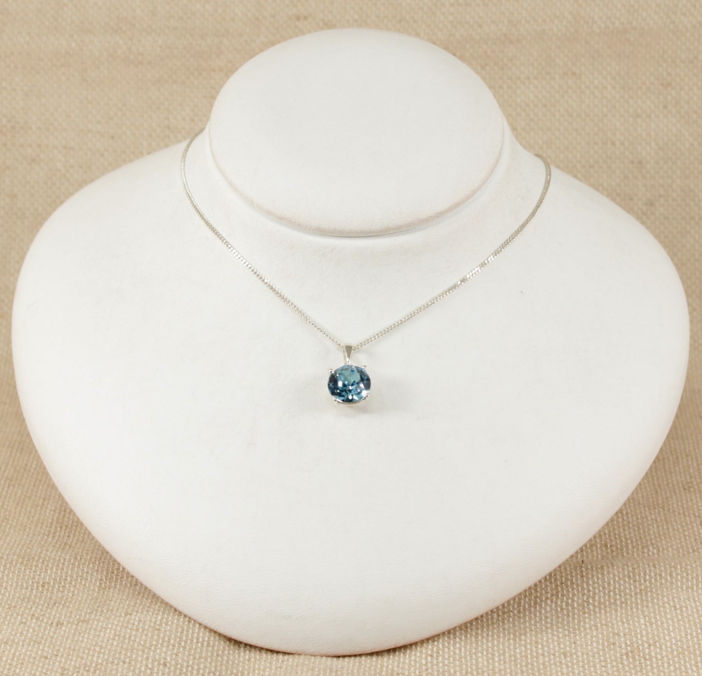 A blue crystal pendant necklace on a silver curb chain, displayed on a white mannequin.