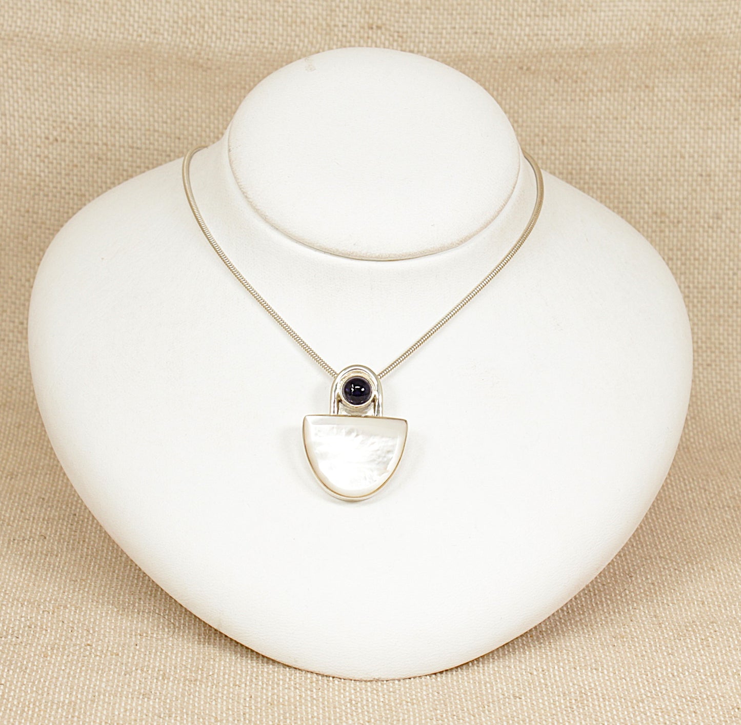 A striking and substantial sterling silver pendant showcasing a sleek, modernist design. The piece features a luminous semicircle of Mother of Pearl, bezel-set to highlight its natural iridescence. Above it rests a round cabochon of deep violet Iolite, adding a touch of rich colour and contrast to the composition.
16 inches long.