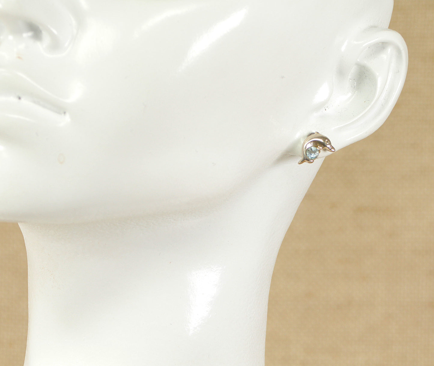 A pair of silver stud earrings in the shape of a dolphin with a round blue topaz placed on a mannequin ear.