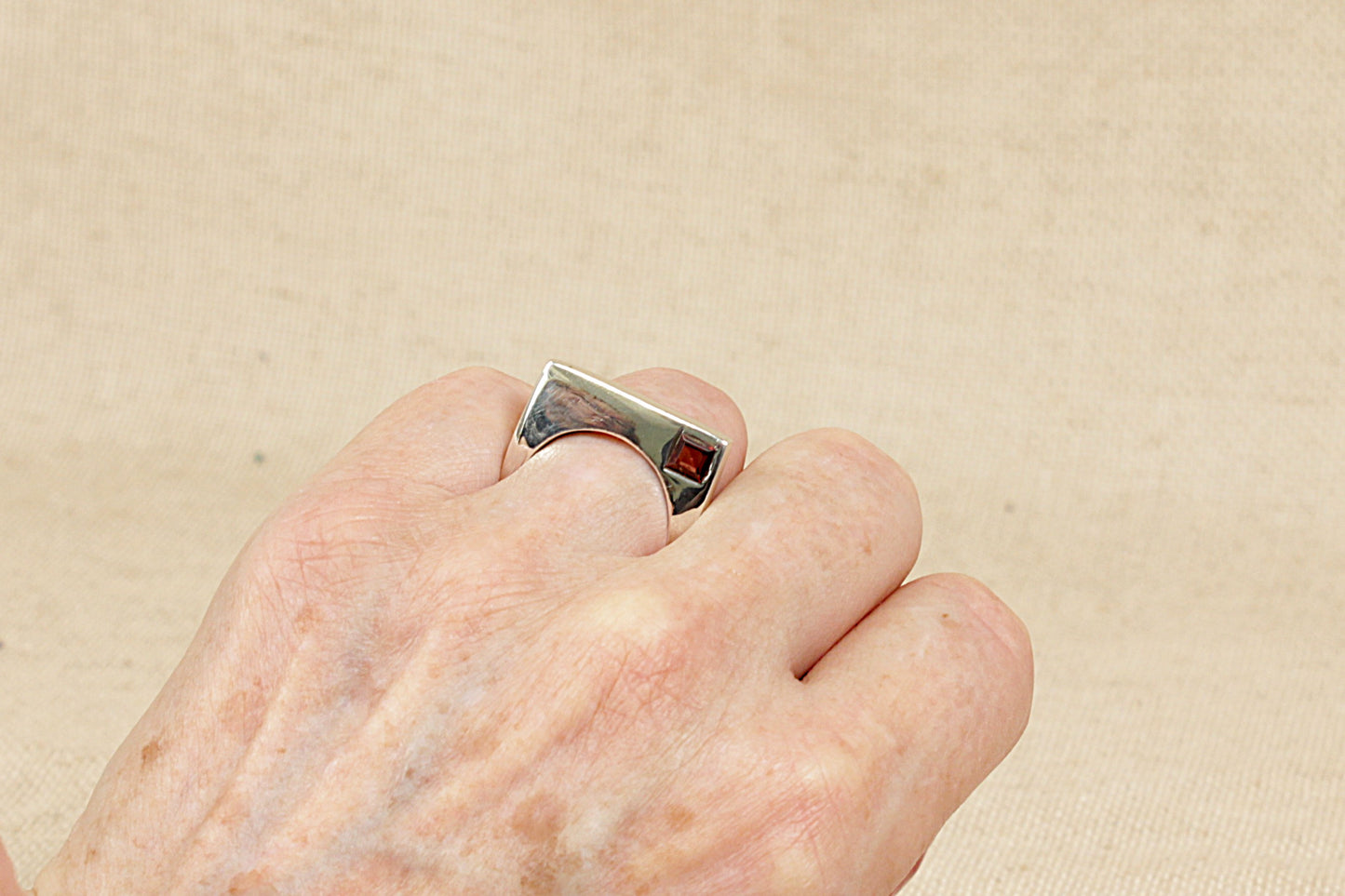 An Interesting Silver and Garnet Modernist Designer Ring.
A striking and contemporary designer sterling silver ring, featuring a unique curved square silhouette with a central round finger cutout for a sculptural, modernist feel. In one corner of the square, a rich square-cut faceted garnet is inset into the silver, adding a bold pop of deep red and a geometric contrast to the fluid shape of the ring.
Minimal yet eye-catching, UK finger size O.