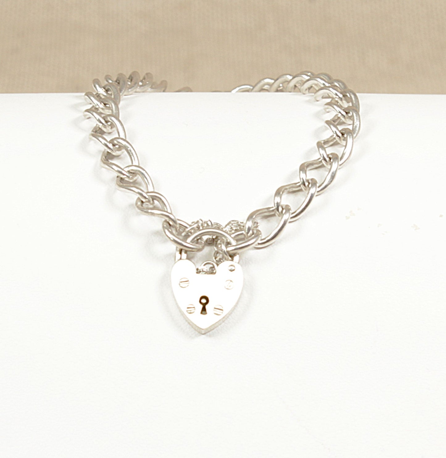 A sterling silver curb link bracelet with a heart-shaped padlock clasp and a safety chain, displayed against a beige background.