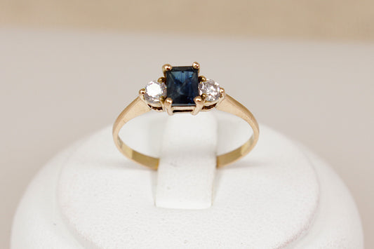 Gold ring with a blue sapphire gemstone and two white cz cubic zirconia gemstones on a white stand.