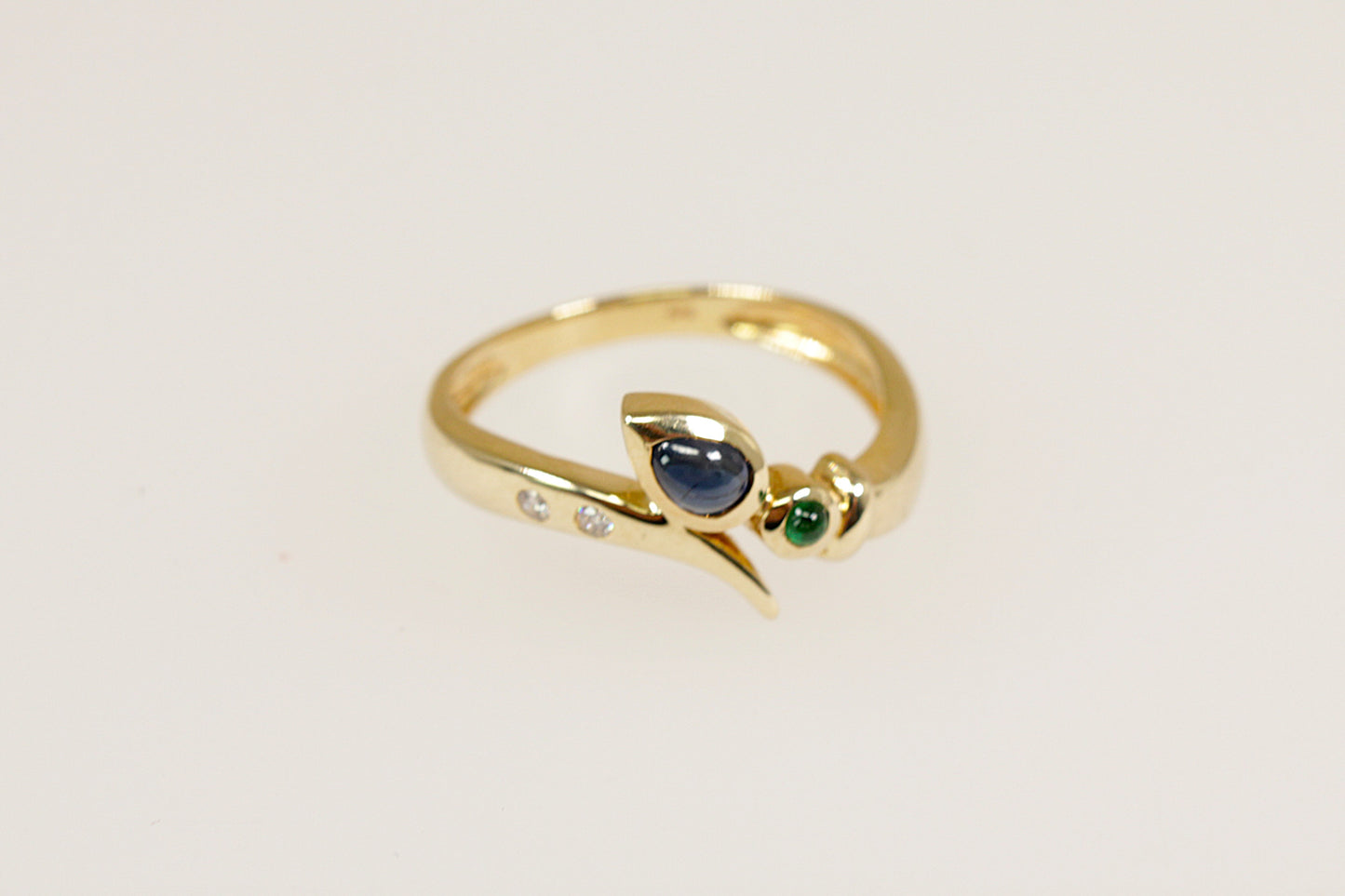 A 14ct gold ring with a teardrop cabochon sapphire, a round cabochon emerald, and two round diamonds set in a flowing twist and crossover setting.