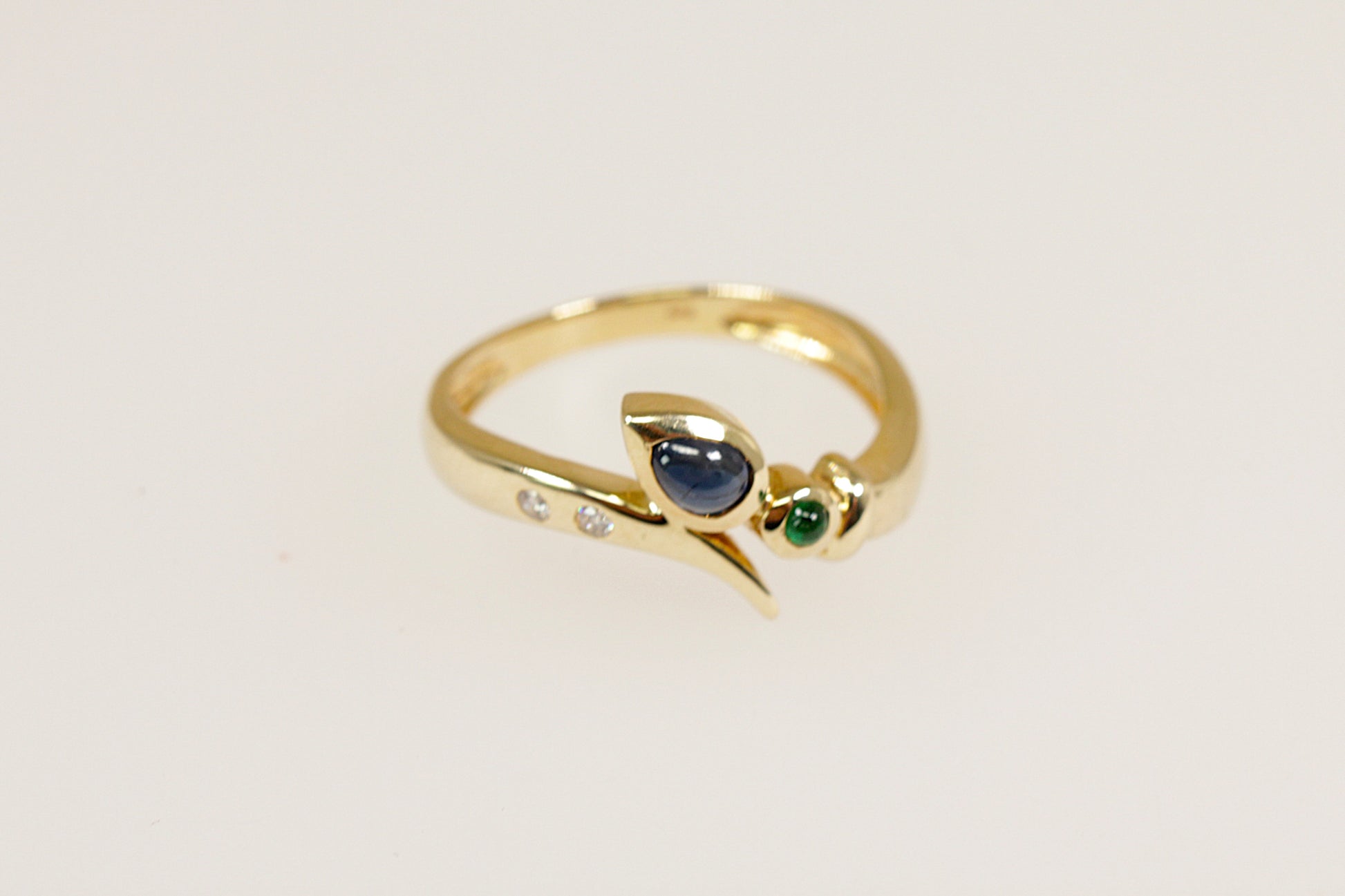 A 14ct gold ring with a teardrop cabochon sapphire, a round cabochon emerald, and two round diamonds set in a flowing twist and crossover setting.