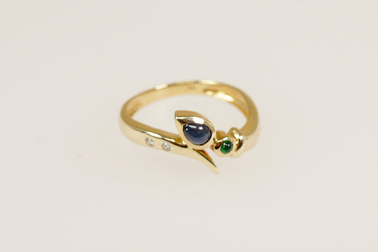 A 14ct gold ring with a teardrop cabochon sapphire, a round cabochon emerald, and two round diamonds set in a flowing twist and crossover setting.