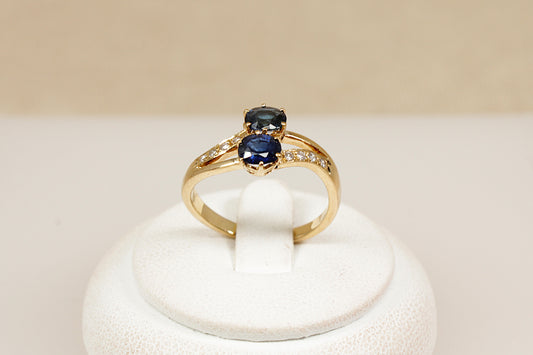 A timeless and romantic design, this 18ct gold ‘Duo’ ring features two round-cut midnight blue sapphires, securely claw-set side by side in a crown-style, split-head setting. The sapphires are positioned on a subtle twist, Each shoulder is adorned with four sparkling diamond accents, adding delicate brilliance and enhancing the ring’s refined elegance. The band is stamped ‘750’ for 18ct gold. UK finger size O.
