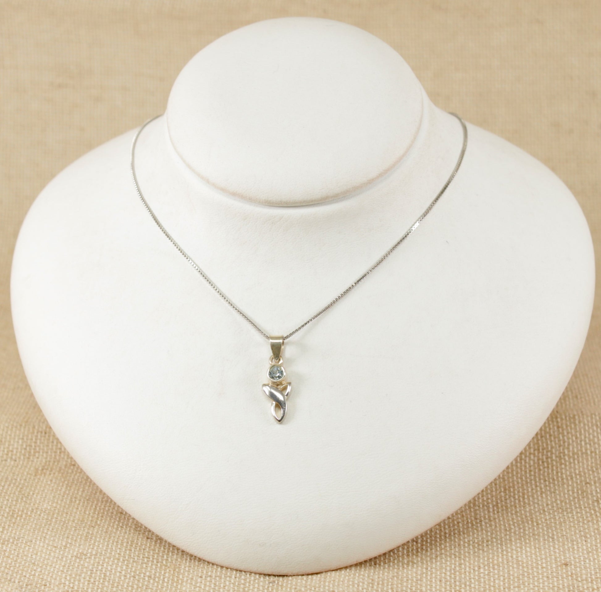 A silver pendant necklace with a round blue topaz set in a Celtic knot design, displayed on a white mannequin.
