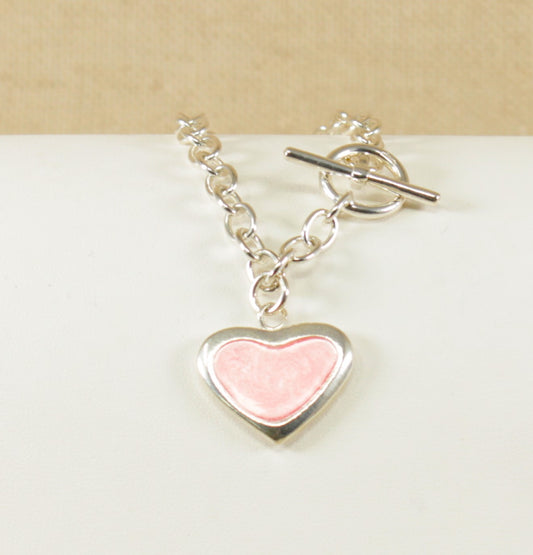 A solid silver bracelet with a belcher design and a 'T' bar fastening, featuring a silver heart charm with pink inlay on a white surface.