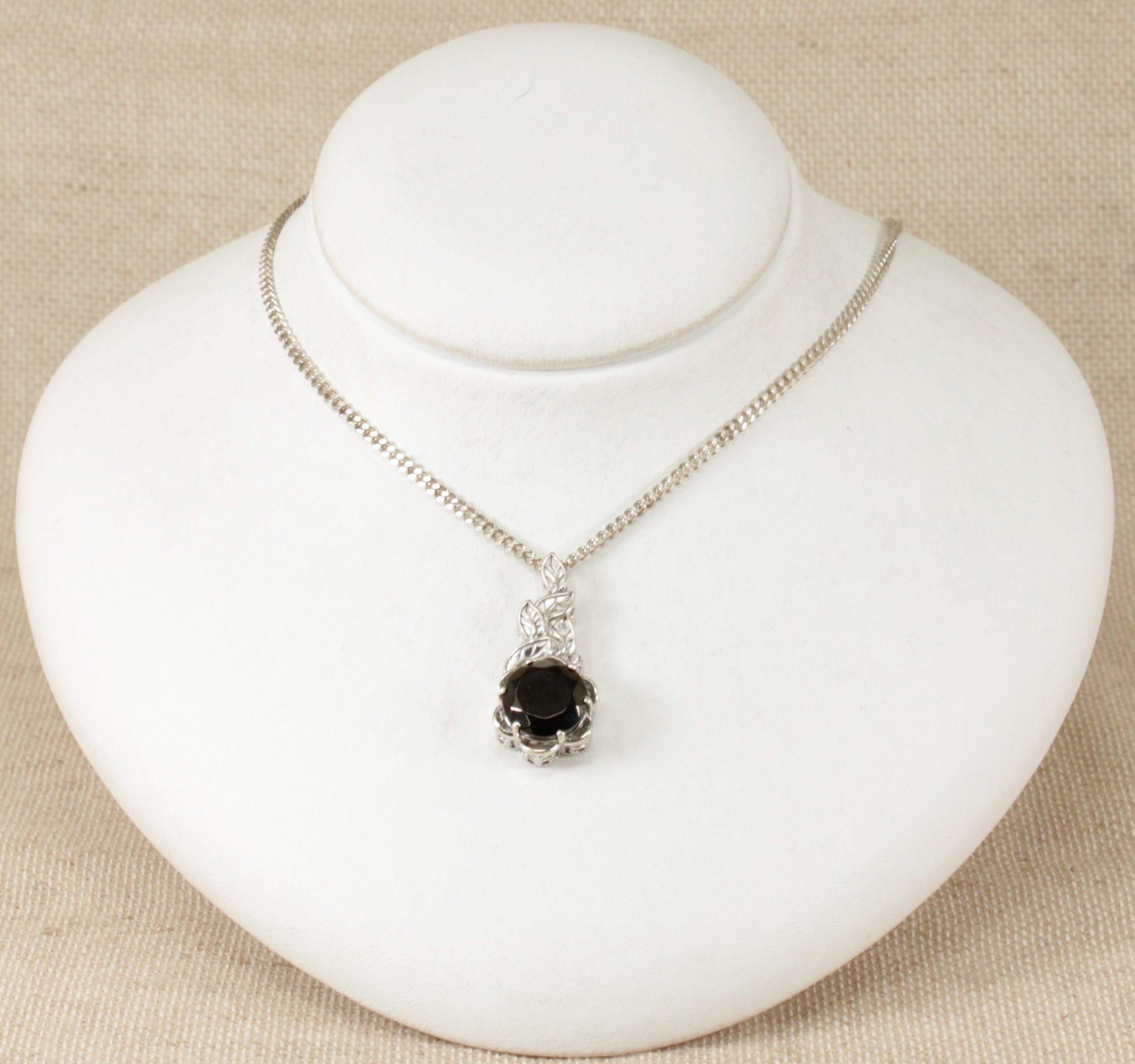 A silver pendant necklace with a round faceted crystal set in a leaf design setting, displayed on a white mannequin.