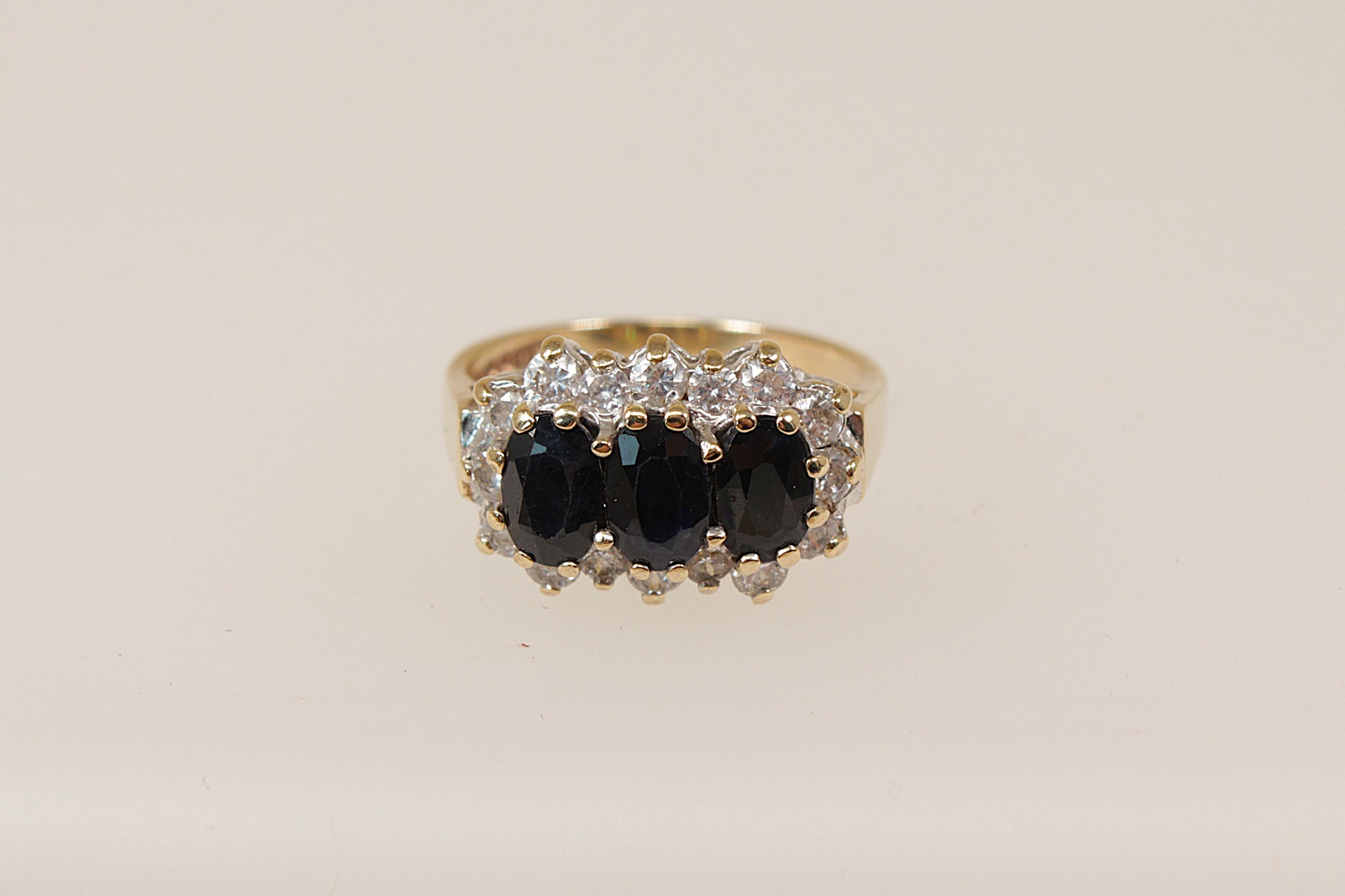 A vintage 9ct gold sapphire trilogy ring with claw set oval blue sapphires and cubic zirconia gemstones around the mount, on a white background.