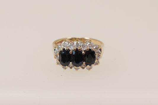 A vintage 9ct gold sapphire trilogy ring with claw set oval blue sapphires and cubic zirconia gemstones around the mount, on a white background.