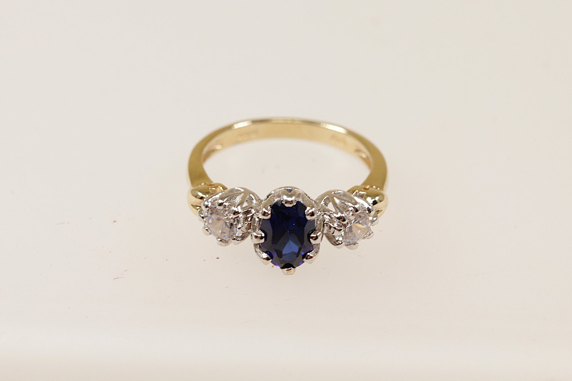 A vintage 9ct gold trilogy ring with a central blue oval-shaped sapphire gemstone and two smaller round cubic zirconia gemstones on either side, set against a white background.