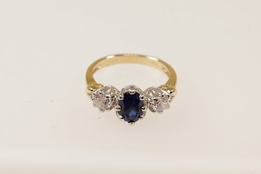 A vintage 9ct gold trilogy ring with a central blue oval-shaped sapphire gemstone and two smaller round cubic zirconia gemstones on either side, set against a white background.