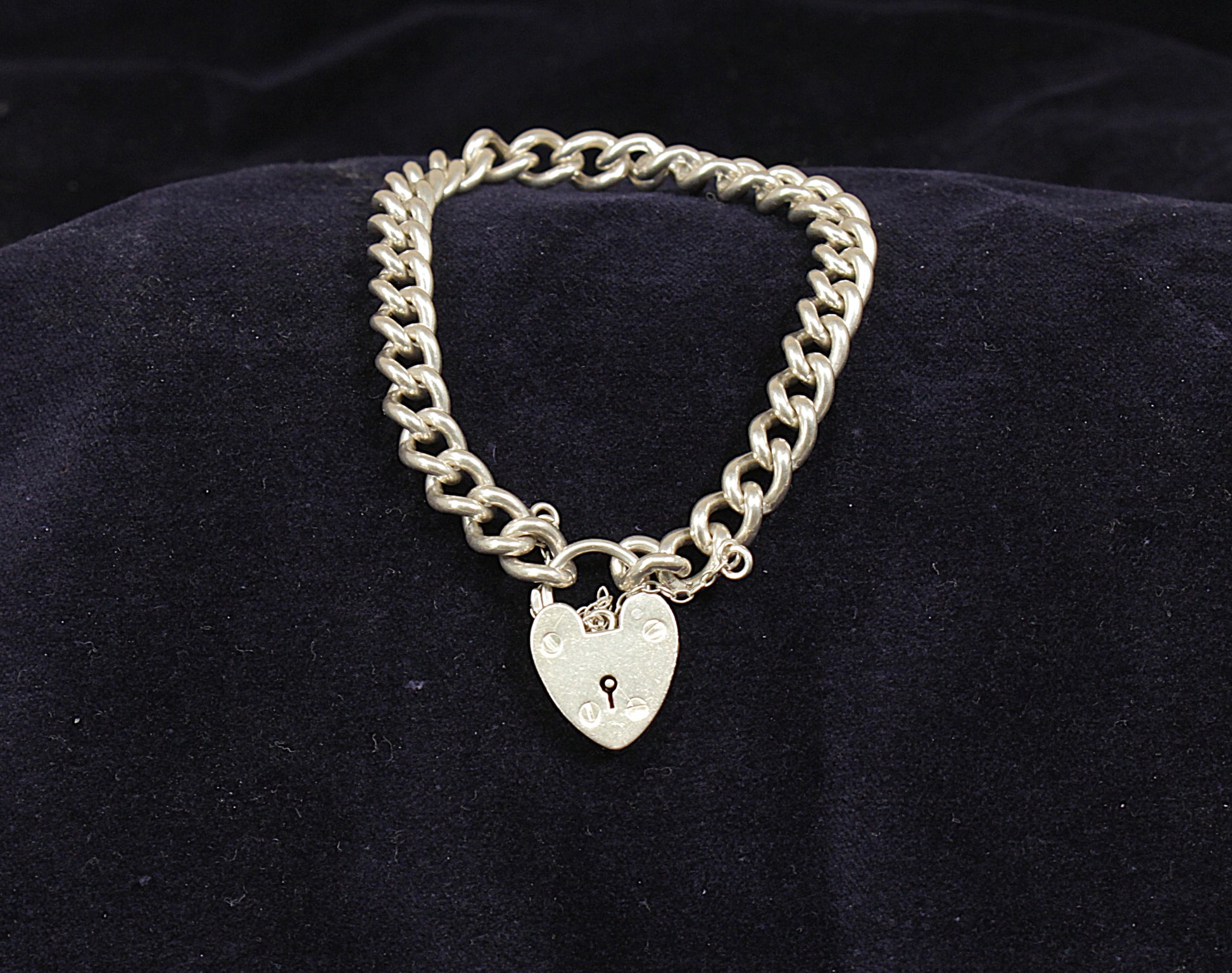 A silver curb link bracelet with a padlock fastening displayed on a dark fabric surface.