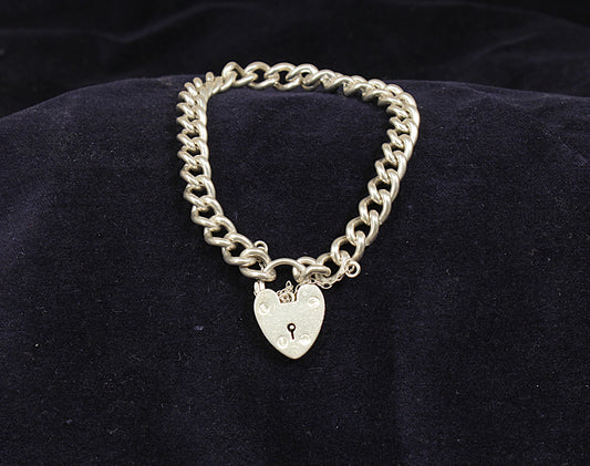 A silver curb link bracelet with a padlock fastening displayed on a dark fabric surface.