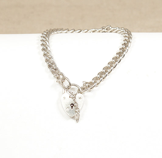A vintage silver curb link bracelet with a heart-shaped padlock clasp and a safety chain, displayed against a beige background.