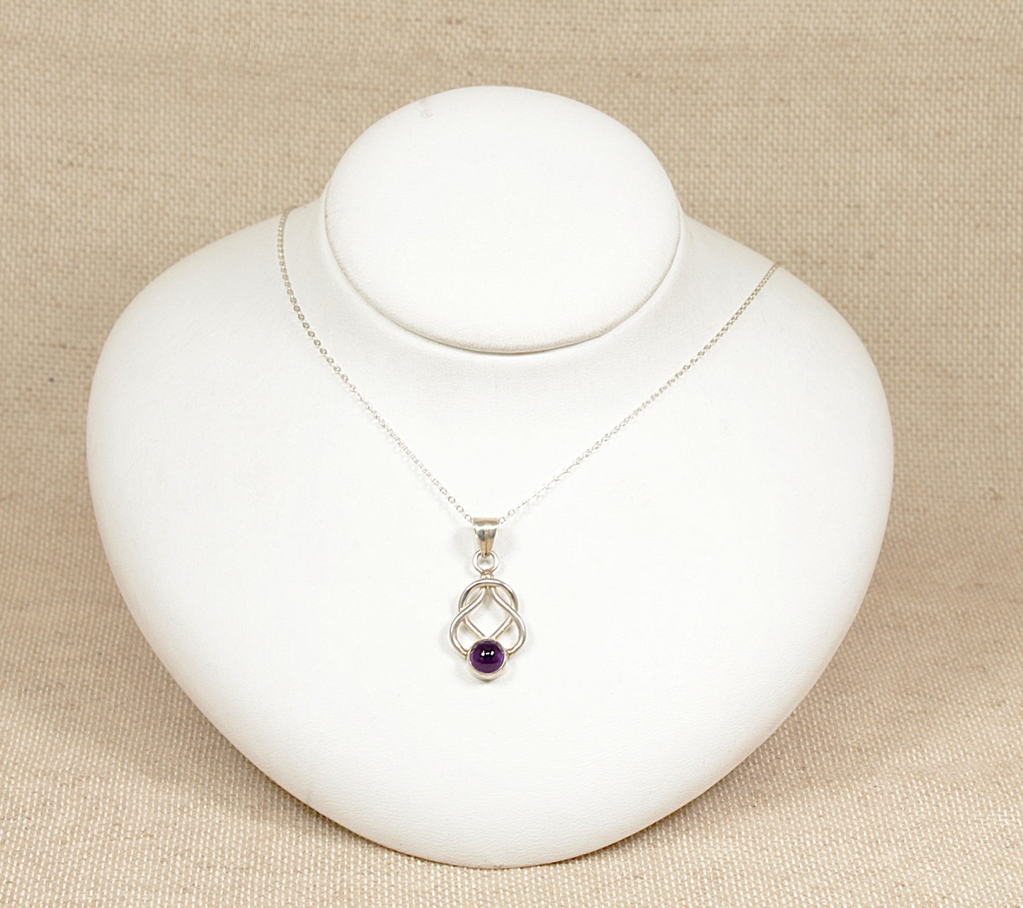 This beautiful and elegant Silver pendant has a timeless and meaningful knot design that suggests unity.
The round cabochon Amethyst at the bottom adds a lovely pop of colour, the bezel set amethyst allows its natural beauty to stand out whilst the bezel setting ensures that the gemstone is secure.
It sits on an 18 inch silver belcher design chain, both are stamped 925 for sterling silver.