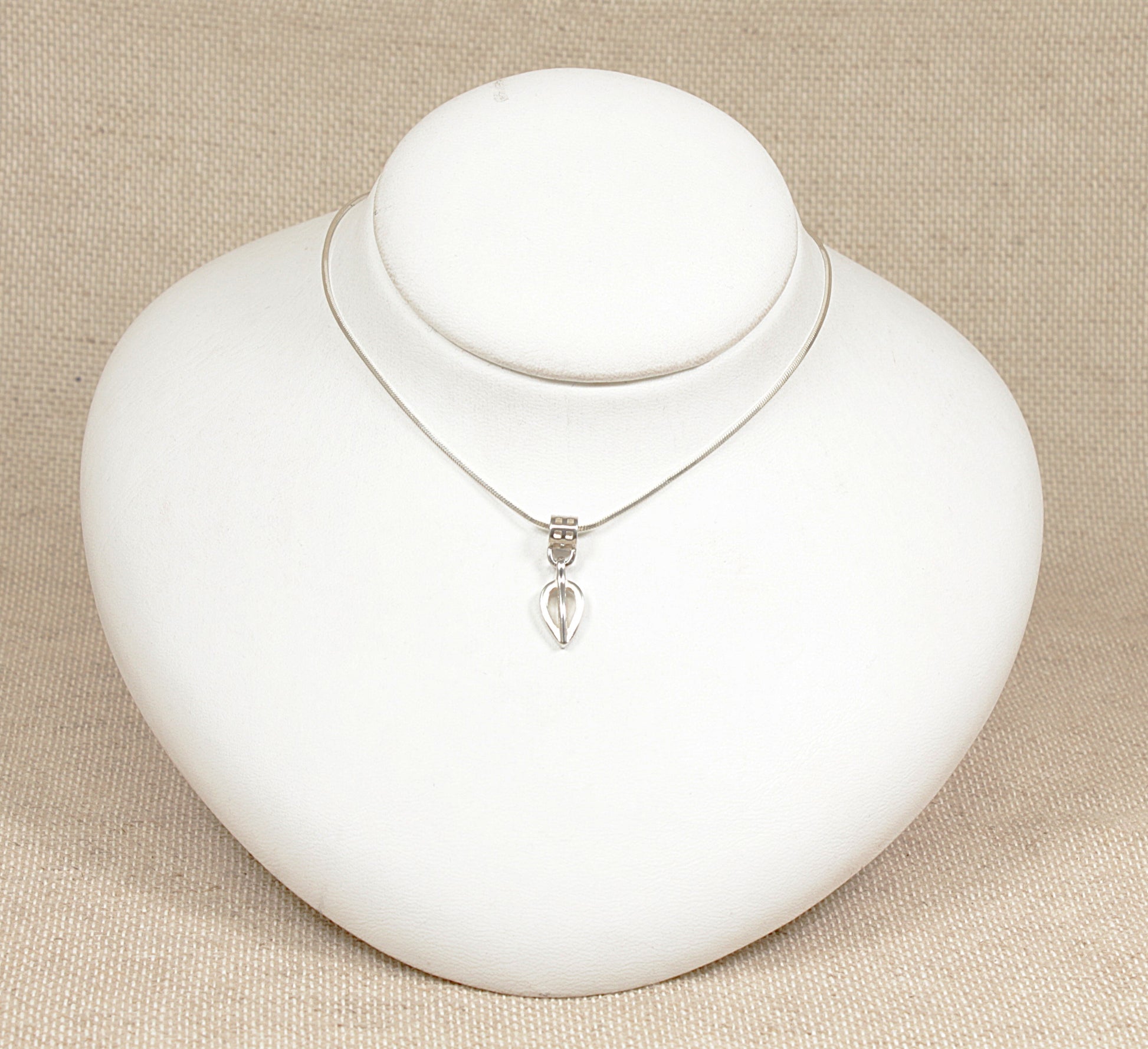 A silver pendant necklace with a geometric and flowing form, displayed on a 16 inch silver snake chain, presented on a white bust against a beige background.