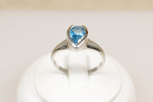 A silver ring with a pear-shaped blue topaz centerpiece set in a bezel, displayed on a white mount.