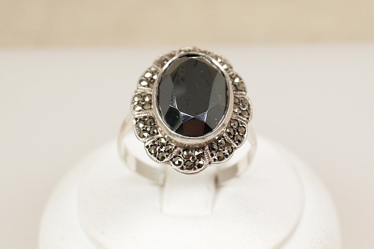 An Art Deco-era sterling silver ring with a large oval faceted hematite stone in a raised bezel setting, surrounded by two rows of marcasite stones, displayed on a white mount.