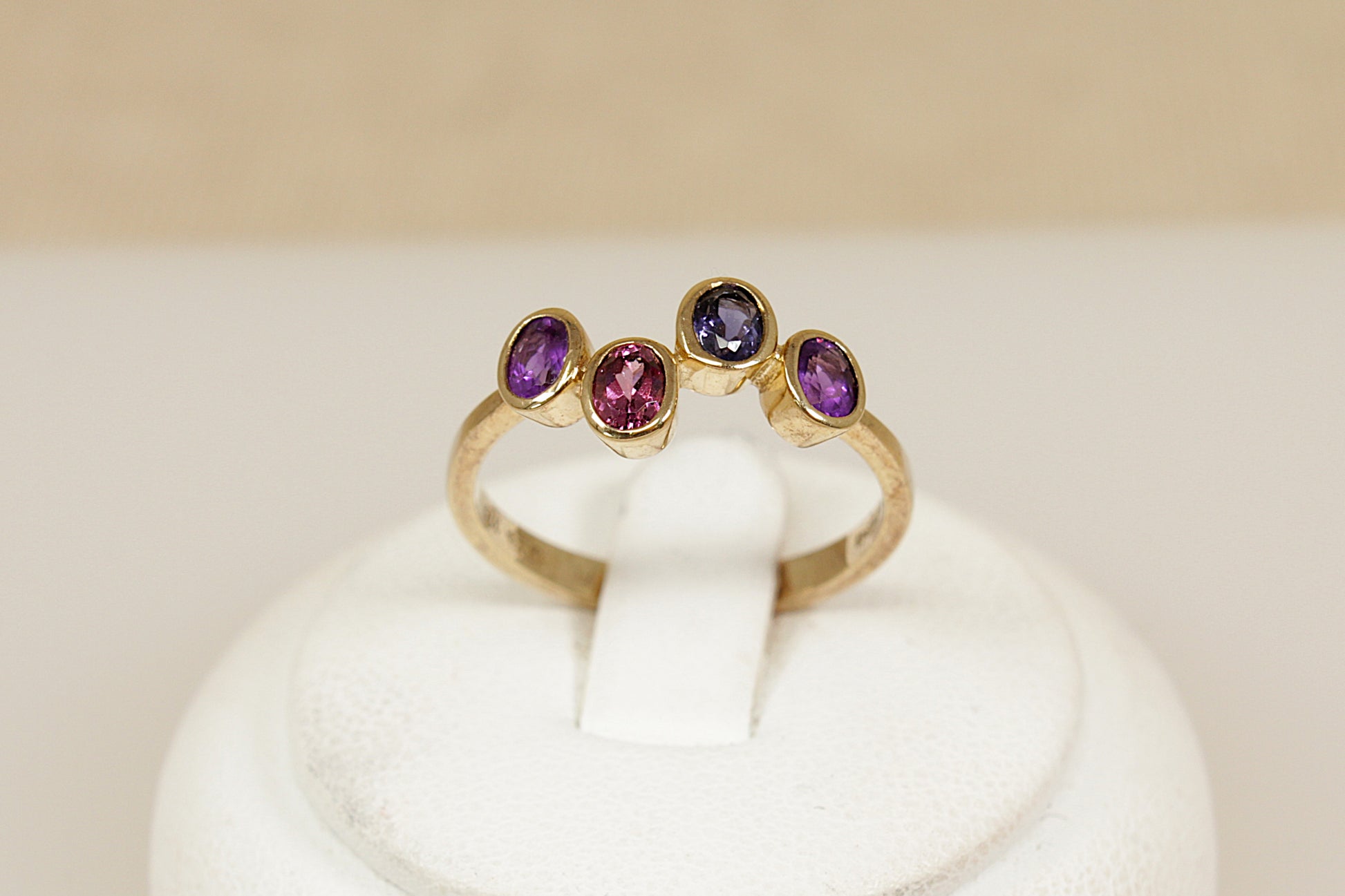 Gold multi gemstone ring with amethyst garnet and blue iolite gemstones on a white stand against a beige background