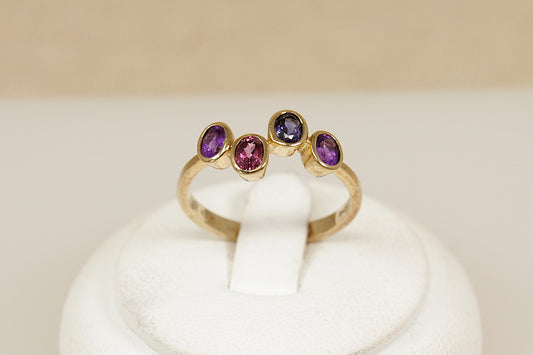 Gold multi gemstone ring with amethyst garnet and blue iolite gemstones on a white stand against a beige background