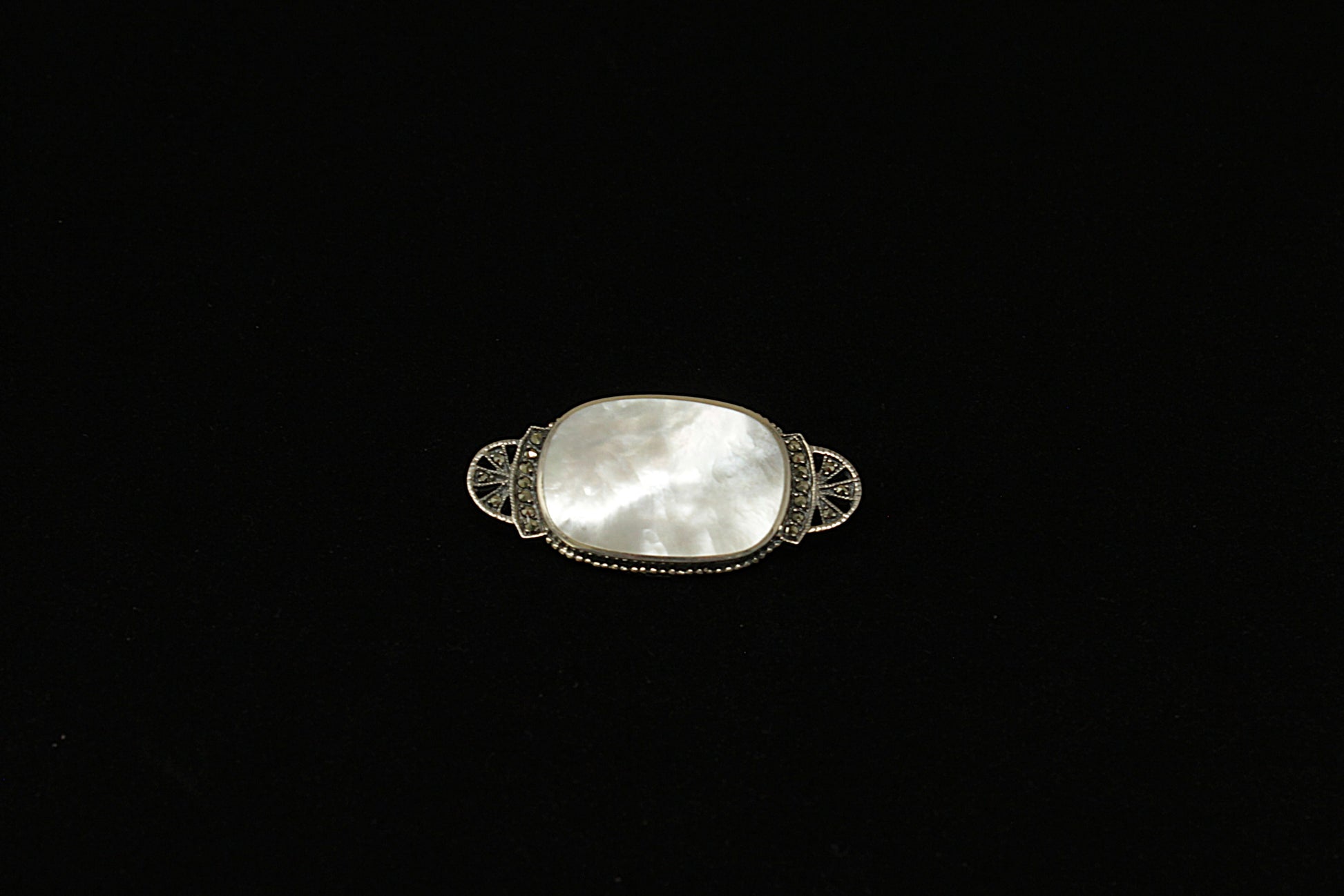 A beautifully crafted Sterling Silver brooch with an elegant Art Deco design, featuring a smooth oval mother of pearl centrepiece that shimmers with subtle iridescence. At each end of the centre MOP is a curved silver band set with glittering marcasite stones, leading into a fan-shaped design adorned with additional marcasite for extra vintage sparkle.
