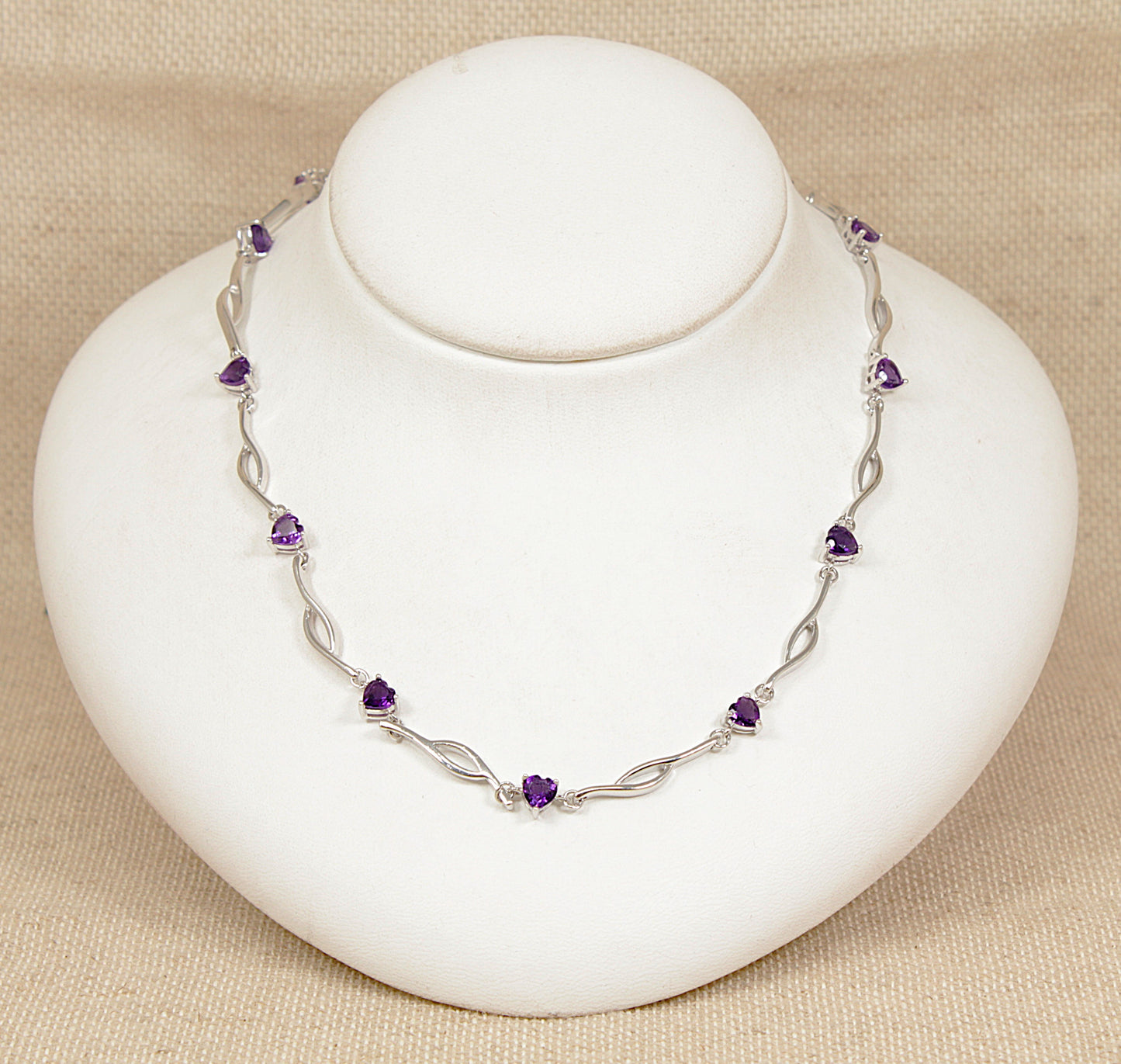 Silver necklace with purple amethyst heart shape gemstones on a white stand against a beige background