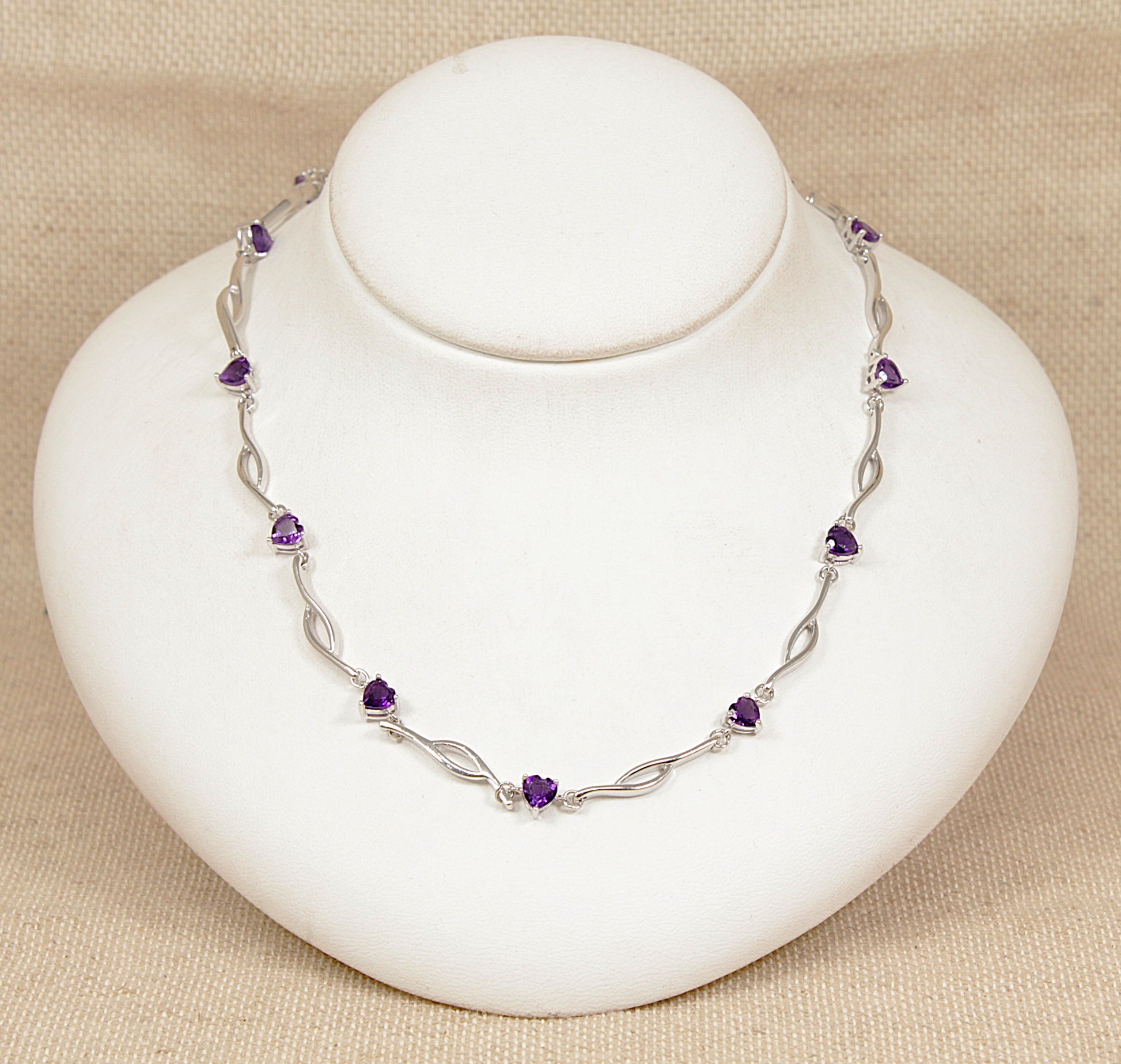 Silver necklace with purple amethyst heart shape gemstones on a white stand against a beige background
