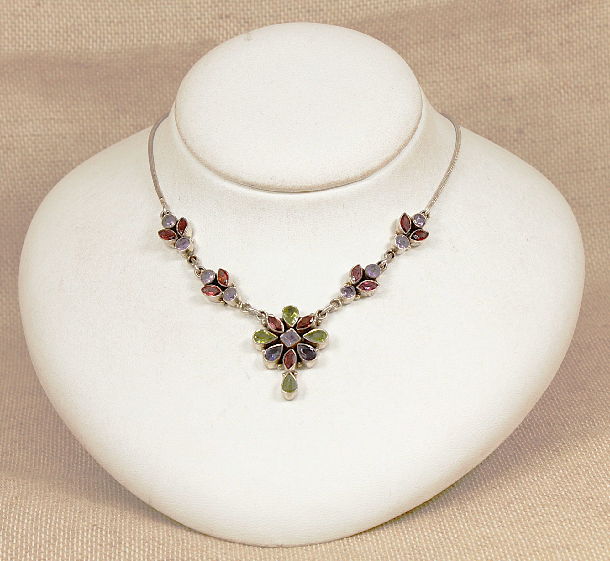 Necklace with multi gemstones peridot, garnet, amethyst in a floral design on a white stand against a beige background