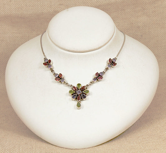 Necklace with multi gemstones peridot, garnet, amethyst in a floral design on a white stand against a beige background