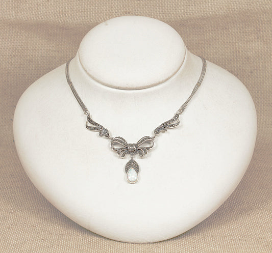 Silver necklace with an opal pendant suspended from a bow pendant encrusted with marcasite on a white stand against a beige background