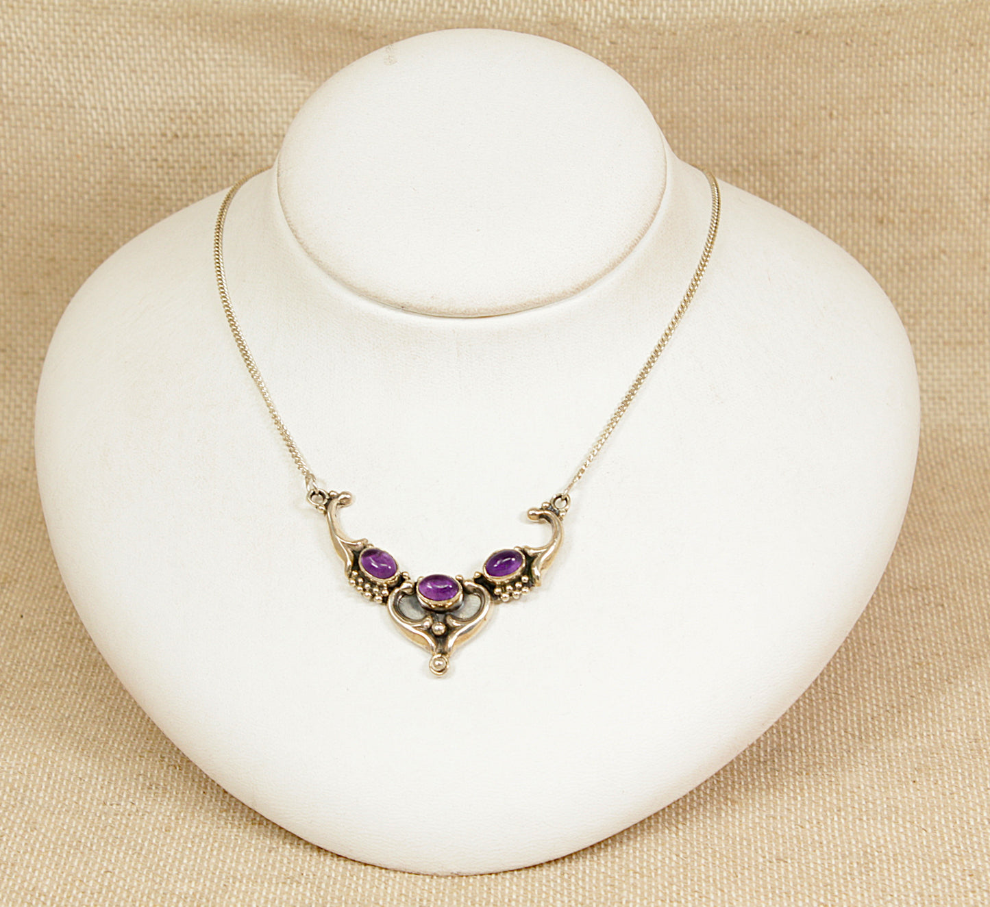 Silver art nouveau necklace with amethyst gemstones on a white stand against a beige background