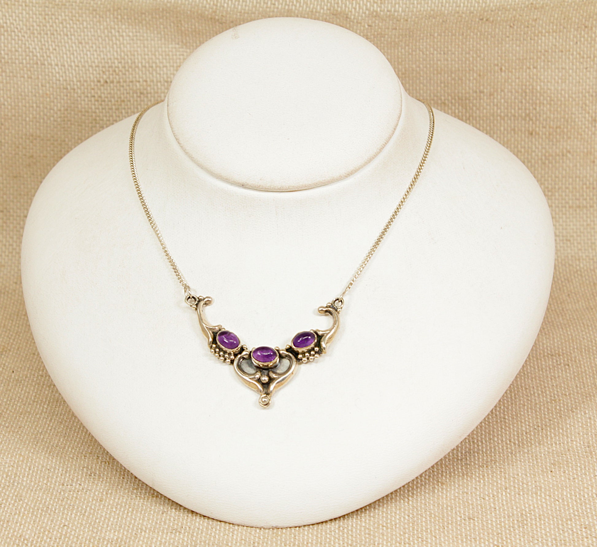 Silver art nouveau necklace with amethyst gemstones on a white stand against a beige background