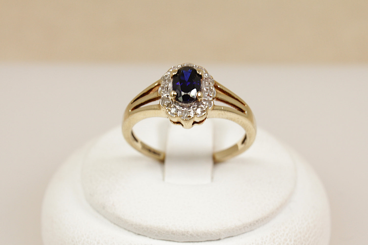 Gold ring with a blue sapphire gemstone and a halo of sparkling diamonds on a white stand against a beige background