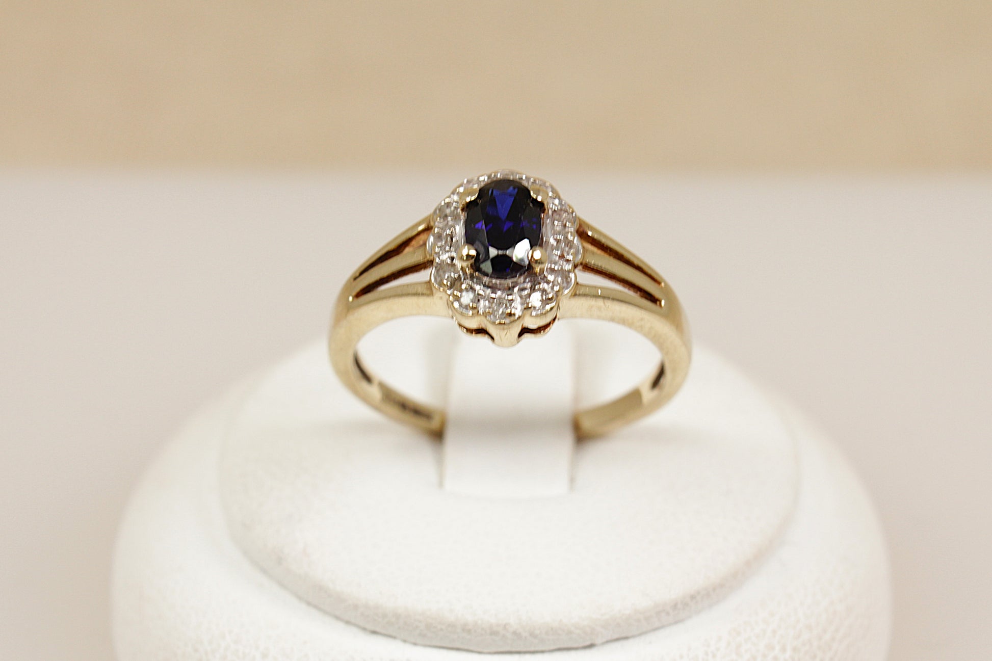 Gold ring with a blue sapphire gemstone and a halo of sparkling diamonds on a white stand against a beige background