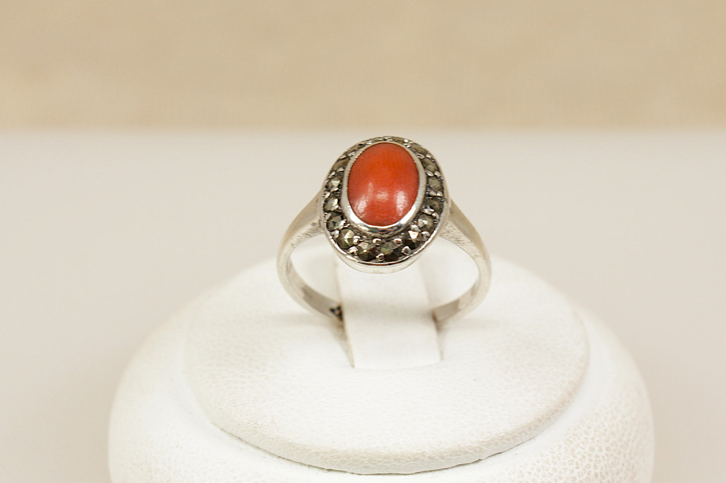 Silver ring with a coral gemstone surrounded by  marcasite gemstones  on a white stand against a beige background.