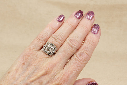 Hand wearing a vintage art deco marcasite silver ring with pink glittery nails on a beige background