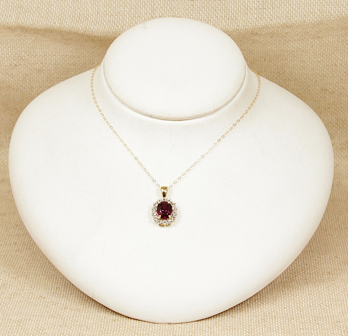 Gold necklace with a red almandine garnet gemstone surrounded by a halo of white topaz gemstones pendant on a white mannequin bust against a beige background