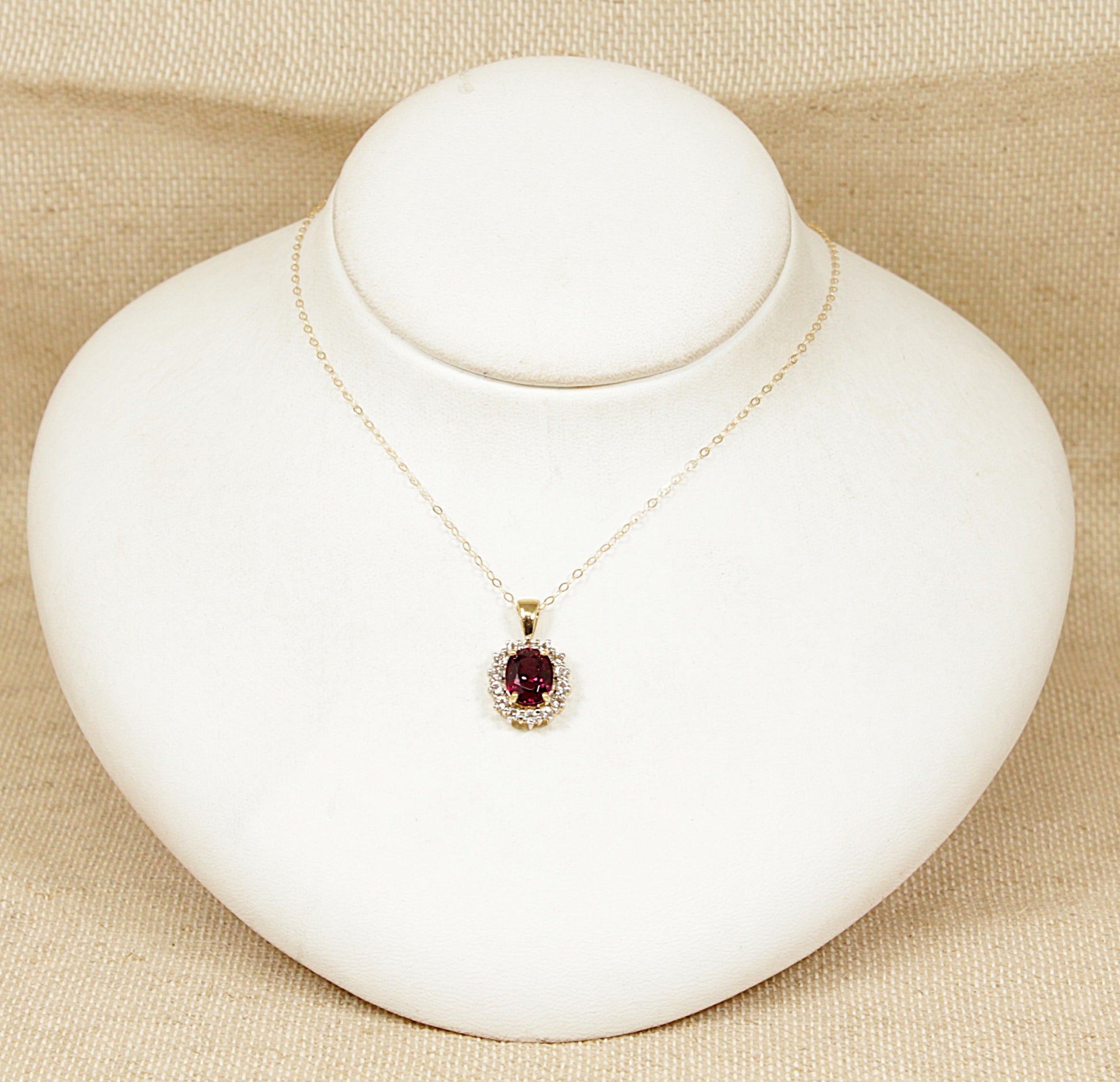 Gold necklace with a red almandine garnet gemstone surrounded by a halo of white topaz gemstones pendant on a white mannequin bust against a beige background
